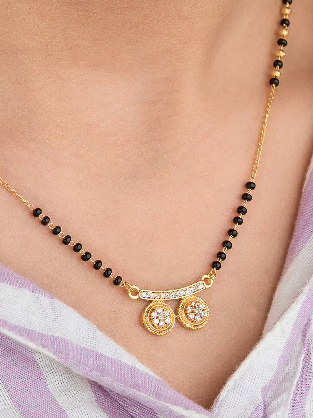 Gold necklace with black beads and circular pendants on a person wearing a light purple garment.