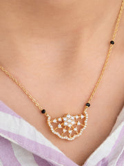Gold necklace with black beads and a decorative pendant on a person wearing a light purple garment.