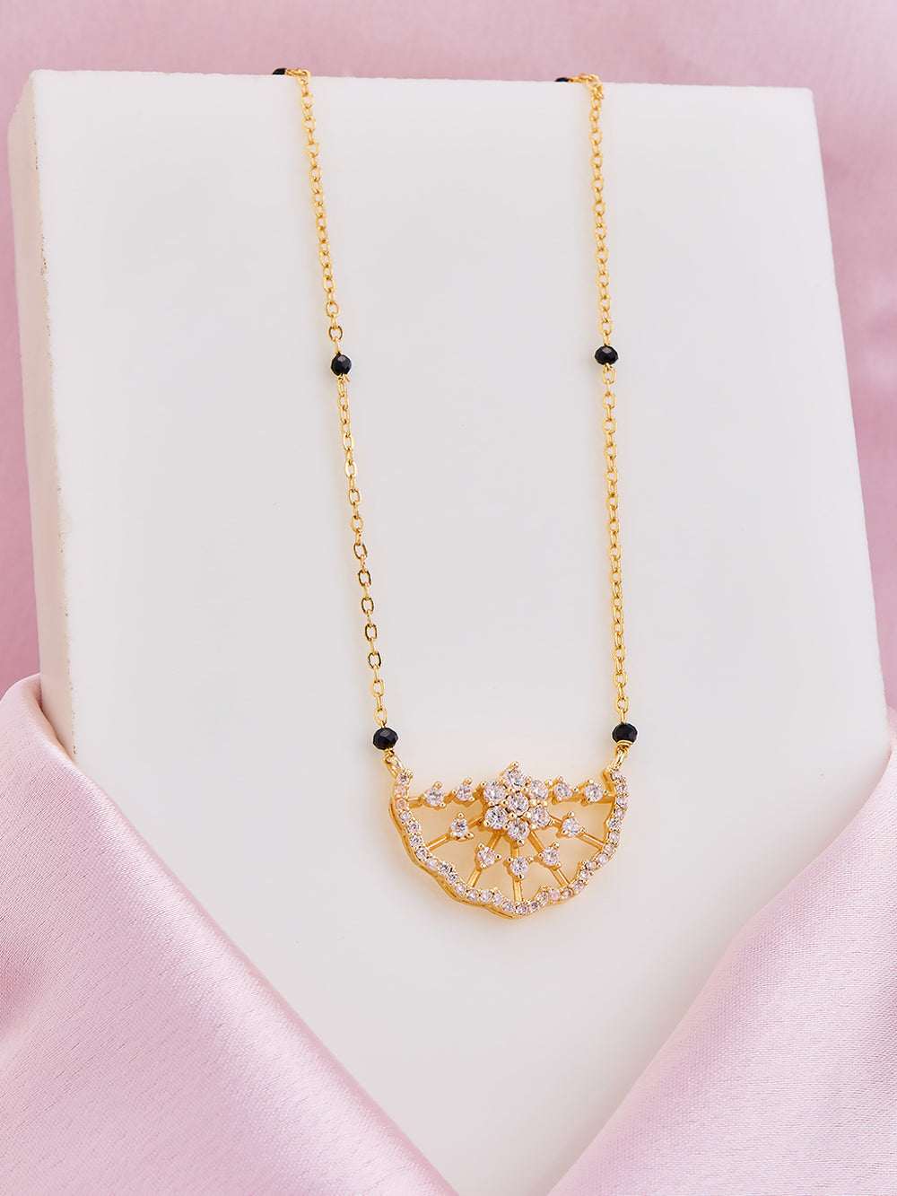 Gold necklace with a floral pendant on a white stand against a pink background