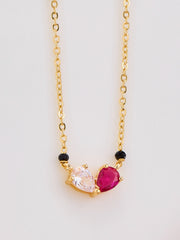 Gold necklace with heart-shaped pink gemstone and black beads on a light gray background