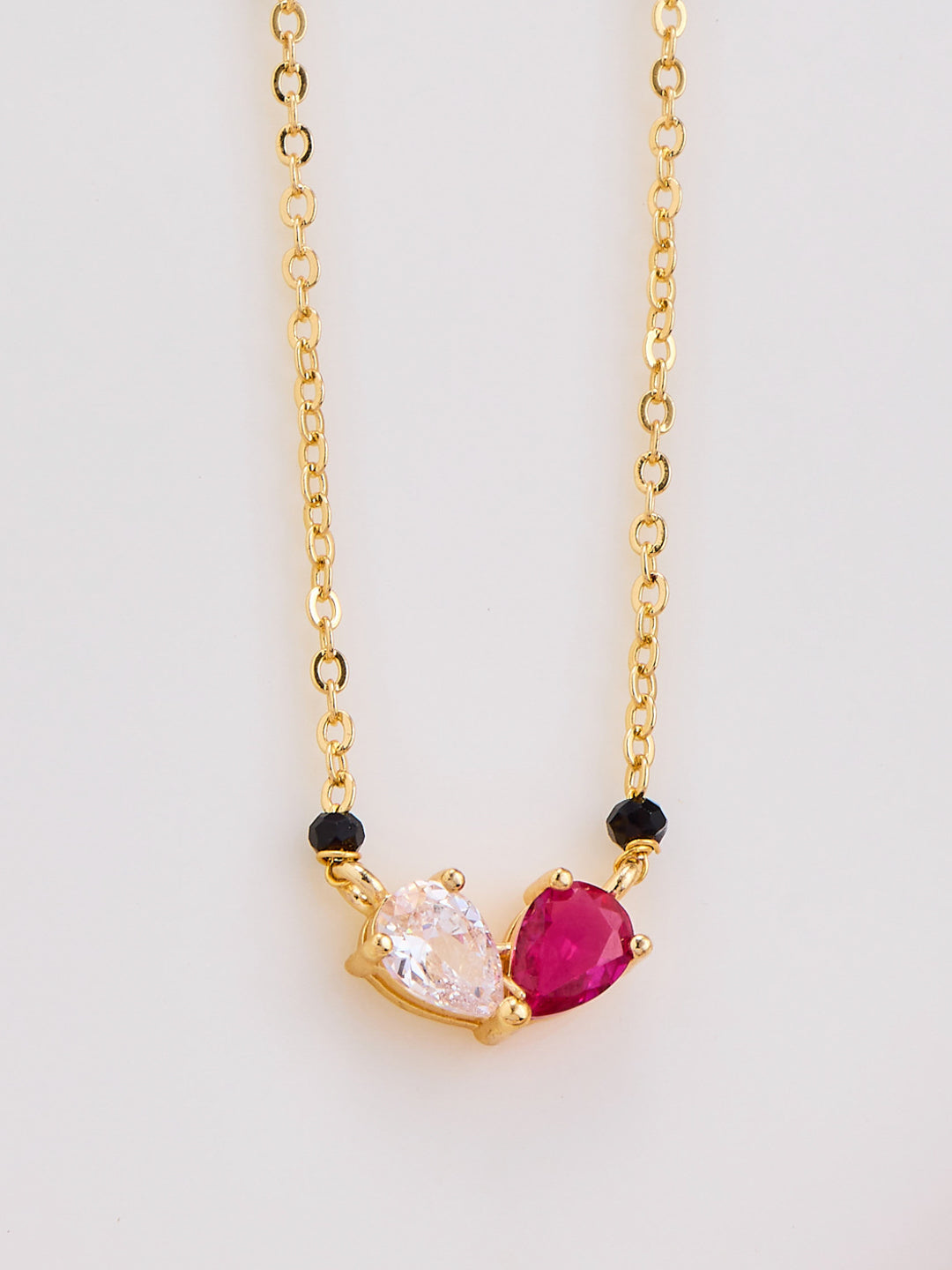 Gold necklace with heart-shaped pink gemstone and black beads on a light gray background