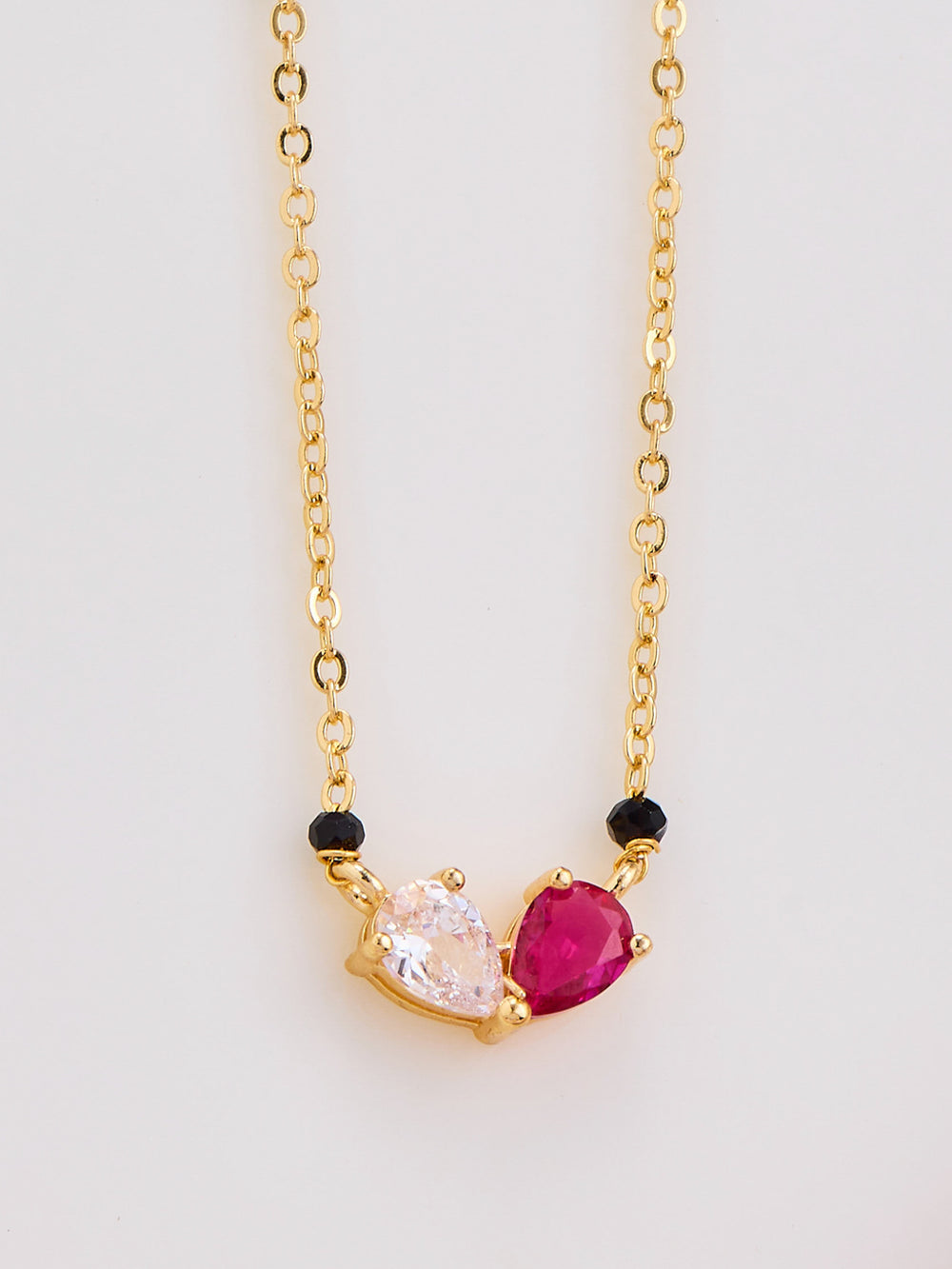Gold necklace with heart-shaped pink gemstone and black beads on a light gray background