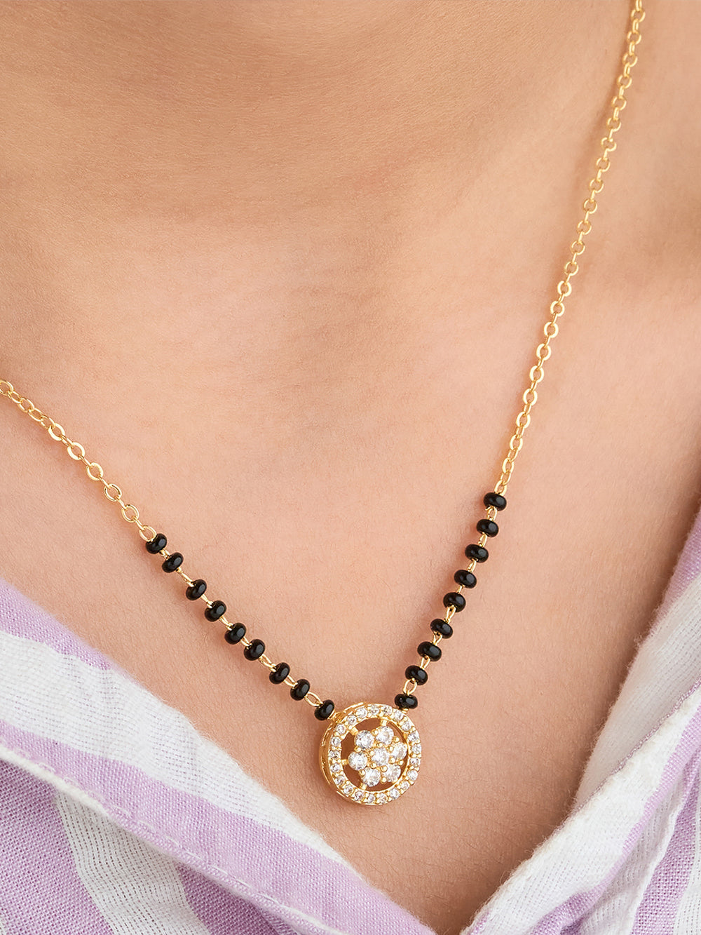 Gold necklace with black beads and a circular pendant on a person wearing a light purple garment.